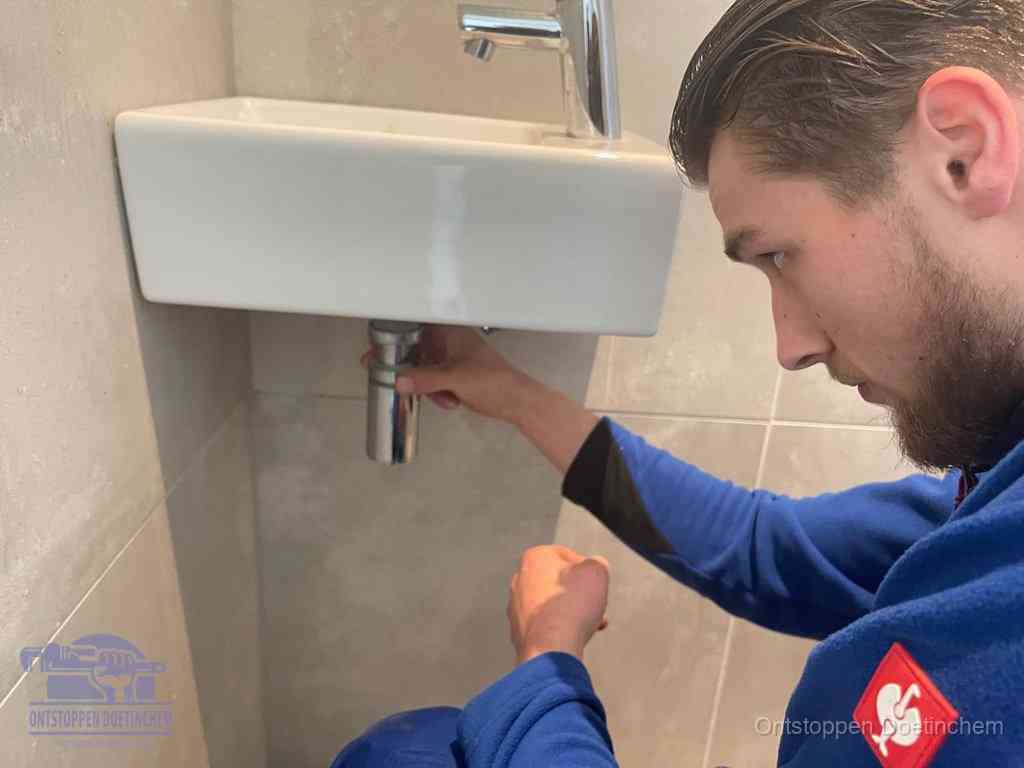 Een monteur draait met de hand de zwanenhalssifon onder een fonteinwasbak in de toiletruimte los om de bron van rioollucht in Doetinchem te vinden